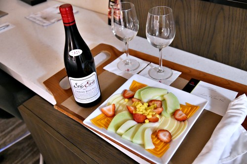 A wooden tray with a bottle of red wine, two glasses, and a fruit platter featuring melon, strawberries, and mango.
