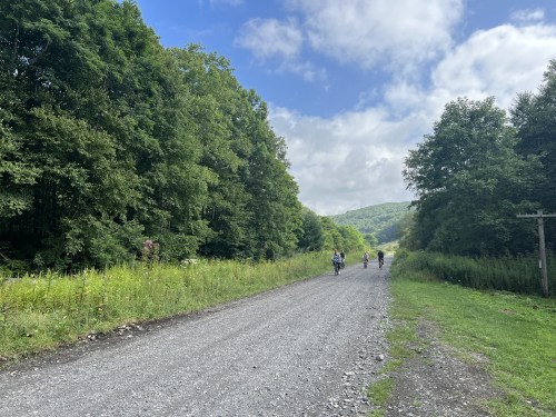 Two cyclists ride along a gravel path surrounded by lush green trees and a blue sky with scattered clouds.