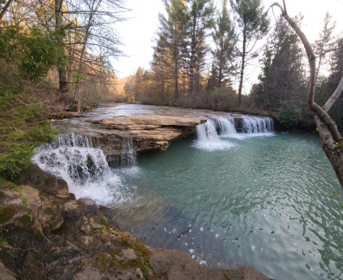 A serene waterfall cascades over rocky ledges into a turquoise pool, surrounded by lush green trees.