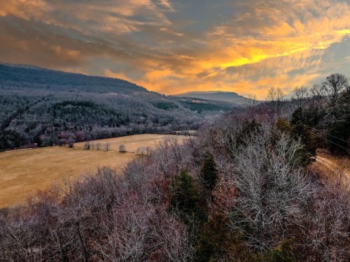 A scenic view of a valley at sunset, with rolling hills, bare trees, and a colorful sky filled with clouds.