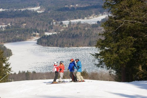 A group of skiers stands on a snowy slope, overlooking a frozen lake and forested landscape in the background.