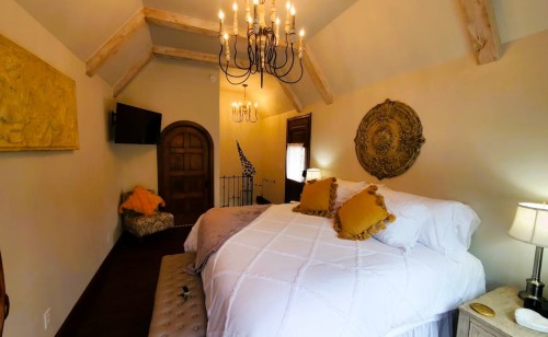 Cozy bedroom with a large bed, decorative pillows, chandelier, and rustic decor, featuring warm tones and wooden accents.