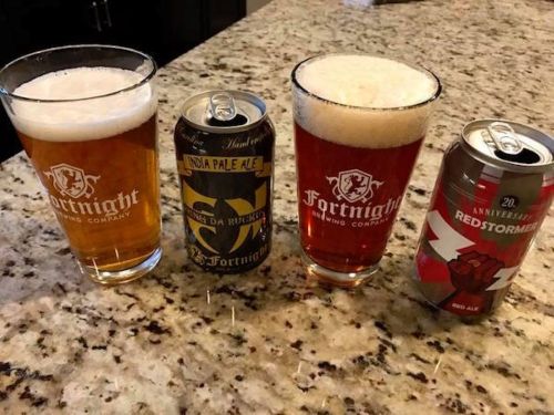 Four beers on a granite countertop: two glasses of Fortnight Brewing Company beer and two cans, including an IPA and a red ale.
