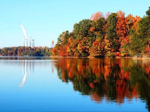 A serene lake reflects vibrant autumn foliage and distant smokestacks under a clear blue sky.