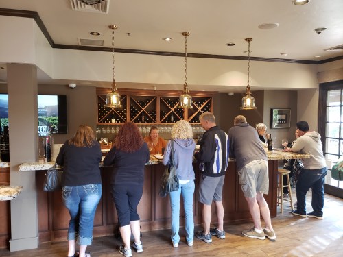 A group of people at a wine tasting bar, chatting with a staff member behind the counter. Wine bottles are displayed on shelves.