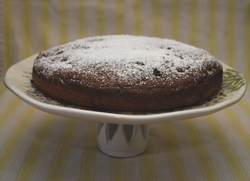 A round chocolate cake dusted with powdered sugar, displayed on a decorative cake stand against a striped background.