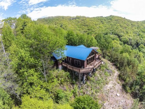 Aerial view of a cabin with a blue roof, surrounded by lush green trees and hills under a partly cloudy sky.