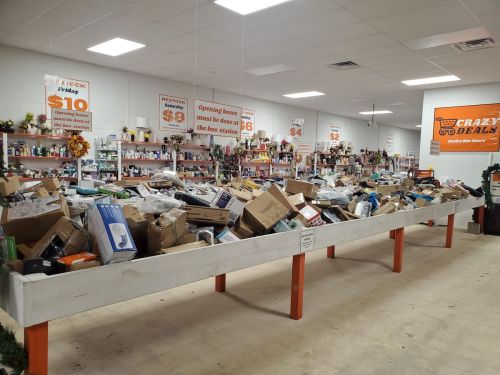 A cluttered store interior with a large table overflowing with boxes and products, surrounded by shelves of items.