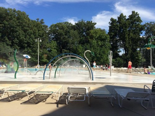 A sunny pool area with water features, lounge chairs, and people enjoying the water amidst green trees.