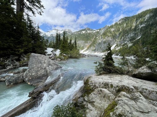 A serene river flows through a mountainous landscape, surrounded by trees and rocky terrain under a partly cloudy sky.