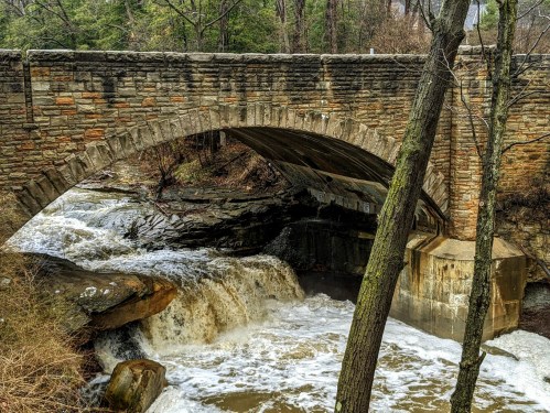 A stone arch bridge spans over a rushing waterfall, surrounded by trees and rocky terrain.