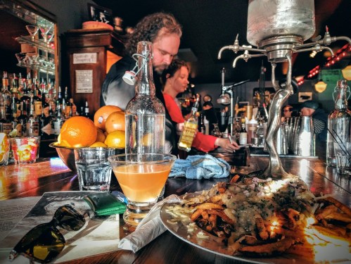 A bar scene featuring a cocktail, fries topped with cheese, and patrons behind the counter with bottles and fruit.