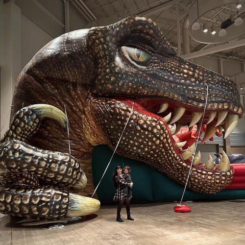 A person holds a child in front of a large inflatable dinosaur with a detailed, menacing face.