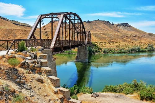 A rusted metal bridge spans a calm river, surrounded by dry hills and blue skies.
