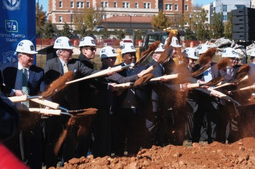 A group of people in hard hats participate in a groundbreaking ceremony, shoveling dirt at a construction site.