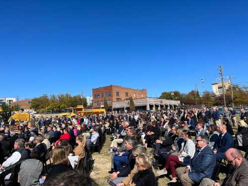 A large crowd gathers outdoors for an event, with buildings and machinery in the background under a clear blue sky.