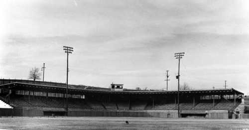 Black and white image of a large, empty stadium with grandstands and floodlights under a cloudy sky.