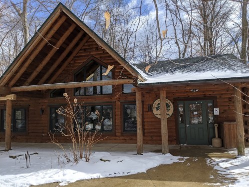 A rustic log cabin with a sloped roof, surrounded by snow and trees, featuring large windows and a welcoming entrance.