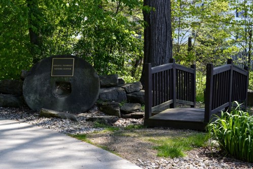 A stone monument with a plaque beside a wooden bridge, surrounded by greenery and a pathway.