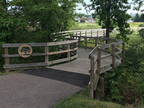 A wooden bridge with a curved design, surrounded by greenery, leading to a pathway in a natural setting.