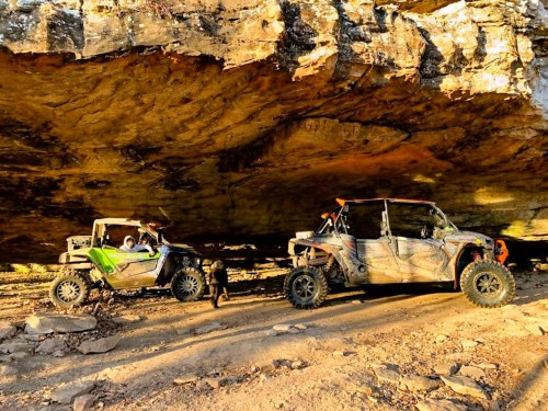 Two off-road vehicles parked under a rocky overhang, with a dog nearby on a dirt path.