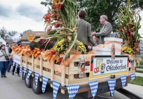 A decorated float with autumn flowers and a "Welcome to Oktoberfest" sign, surrounded by people at a parade.