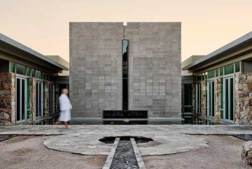 A serene architectural space with a stone wall, water features, and a person in a robe walking through the courtyard.