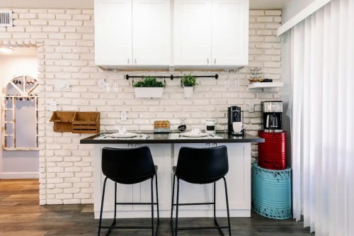 Modern kitchen with white brick walls, black bar stools, and a coffee station, featuring plants and decorative storage.