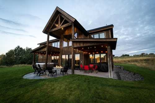 A modern wooden house with large windows, outdoor seating, and a grassy yard under a cloudy sky.