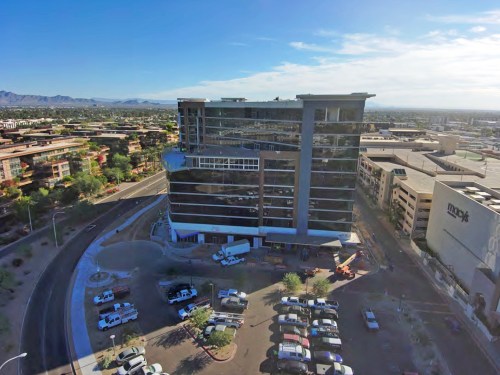 Aerial view of a modern multi-story building surrounded by parking and urban landscape under a clear blue sky.