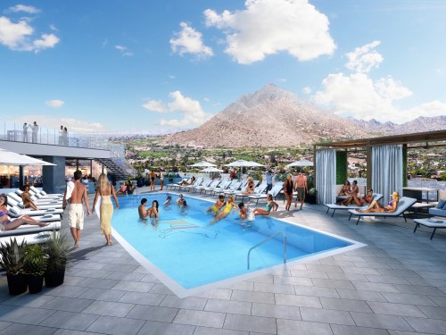 A vibrant pool area with sun loungers, people relaxing, and a mountain backdrop under a clear blue sky.