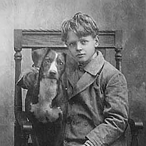 A young boy in a coat sits on a chair with a dog resting beside him, both looking directly at the camera.