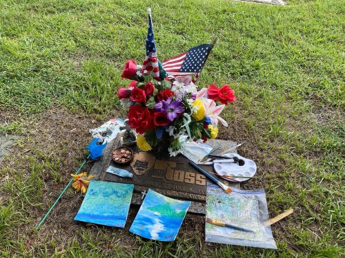 A grave marker surrounded by flowers, an American flag, and paintings on the grass.