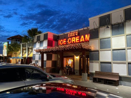 A brightly lit ice cream shop with a sign reading "Kirk's Ice Cream" at dusk, surrounded by palm trees and outdoor seating.