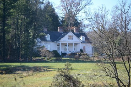 A white house with a porch, surrounded by trees and a grassy field on a clear day.