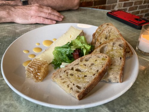 A plate with cheese, honeycomb, salad, and slices of bread, with a hand and a phone in the background.