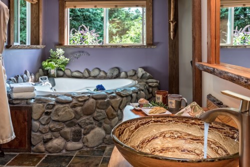 A serene bathroom featuring a stone tub, wooden accents, and a stylish sink with flowing water, surrounded by greenery.