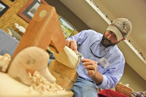 A man carves wood at a workshop, focused on his work with tools and wood shavings around him.
