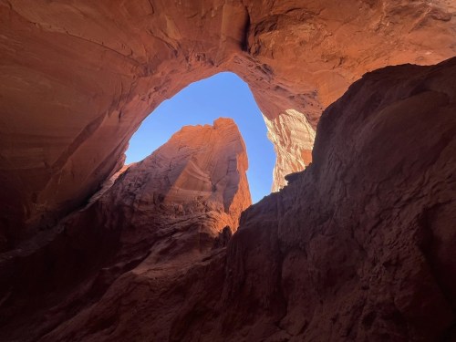 View of a natural rock arch framed by towering red rock formations and a clear blue sky above.