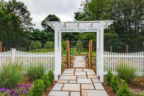 Entrance to the Grain House Organic Garden, featuring a white picket fence and stone pathway surrounded by greenery.
