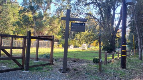 A wooden sign marks the entrance to a grassy area with trees, near a fenced enclosure and power pole.