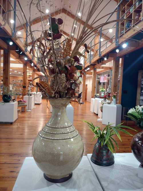 A large decorative vase with dried flowers, set in a spacious gallery with wooden beams and display stands.