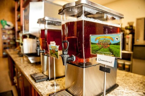 Two beverage dispensers filled with iced tea, labeled "Unsweetened," on a granite countertop in a cafe setting.