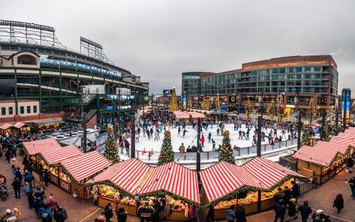 A festive ice skating rink surrounded by holiday market stalls and decorated buildings, with a crowd enjoying the scene.