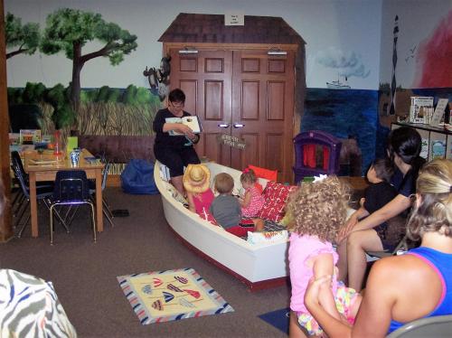 A storyteller reads to children seated in a boat-themed area, surrounded by colorful murals and a cozy atmosphere.