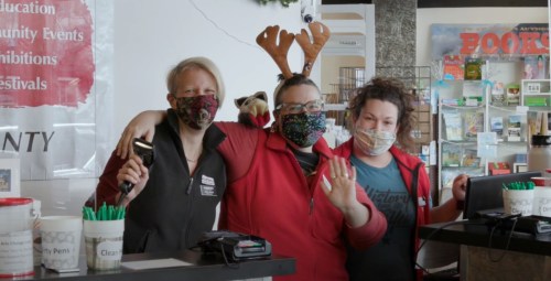 Three smiling staff members wearing masks and festive attire pose together at a counter, with holiday decorations in the background.