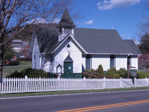 A small white church with a steeple, surrounded by greenery and a white picket fence, set against a blue sky.