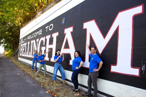 A group of five people in blue shirts poses playfully by a large "Welcome to Bellingham" mural on a wall.