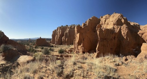 A panoramic view of rugged red rock formations under a clear blue sky, with sparse vegetation in the foreground.