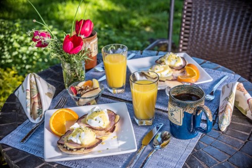 A table set outdoors with two plates of eggs Benedict, orange slices, juice, and coffee, surrounded by flowers.